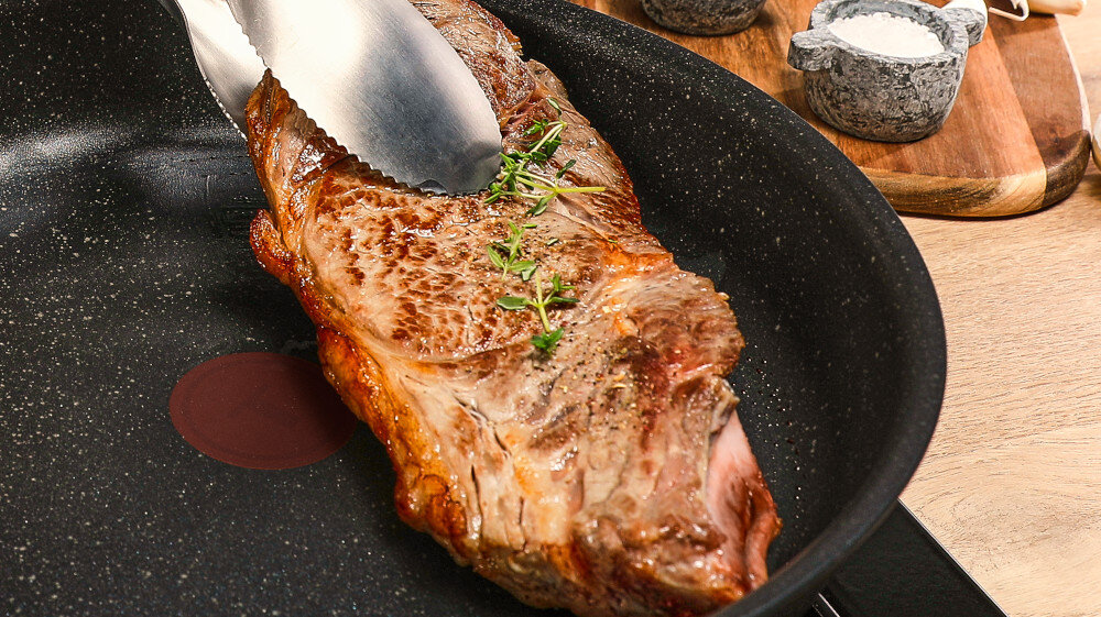 Na patelni znajduje się smażony kawałek mięsa z ziołami, trzymany przez metalowe szczypce. W tle widoczne są dwa kamienne naczynia na drewnianej desce.