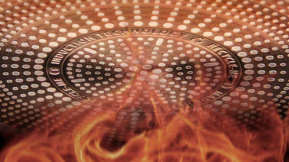 Zdjęcie przedstawia spód patelni z widocznymi okrągłymi wzorami i tekstem \'INDUCTION TECHNOLOGY\'. W dolnej części obrazu widoczne są płomienie.