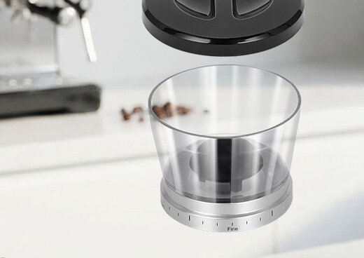 Młynek MMK-09M z przezroczystymi ściankami, nad nim czarna pokrywa, w tle niewyraźny blat kuchenny.