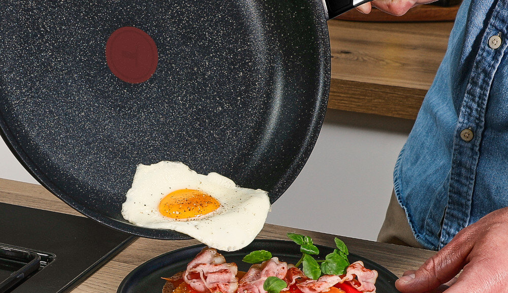 Osoba trzyma patelnię z nieprzywierającą powłoką, na której znajduje się smażone jajko. Na talerzu poniżej leży bekon i zielone liście.