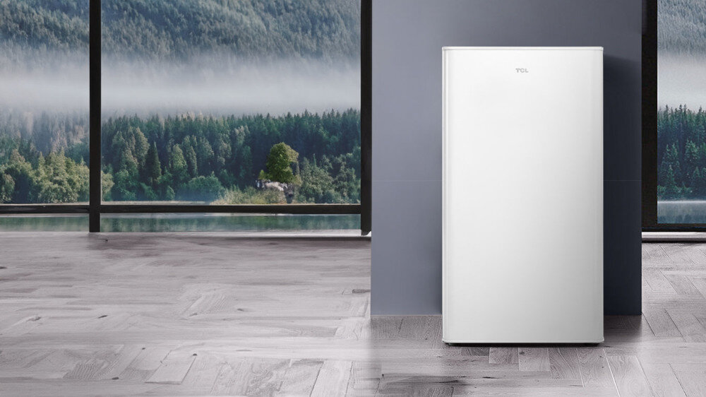 LODÓWKA TCL RF092DWE0 Na grafice RF092DWE0_lifestyle_1.jpg widać białą lodówkę wolnostojącą TCL ustawioną przy gładkiej, szarej ścianie. W tle znajduje się duże okno sięgające od podłogi do sufitu z widokiem na jezioro i zalesione wzgórza, a całość dopełnia jasna podłoga z drewnianych paneli.