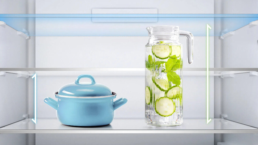 LODÓWKA TCL RF092DWE0 Na zdjęciu przedstawiono wnętrze lodówki z dwiema szklanymi półkami, na których stoją garnek z niebieską pokrywką oraz wysoki dzbanek z wodą, plasterkami ogórka i listkami mięty. Wokół półek zaznaczono kolorowe linie i strzałki w pionie, które sugerują możliwość regulacji wysokości lub zwiększoną przestrzeń na wyższe naczynia regulowane półki elastyczna organizacja wnętrza zmiana wysokości półek miejsce na duże produkty wygodne przechowywanie dopasowanie przestrzeni