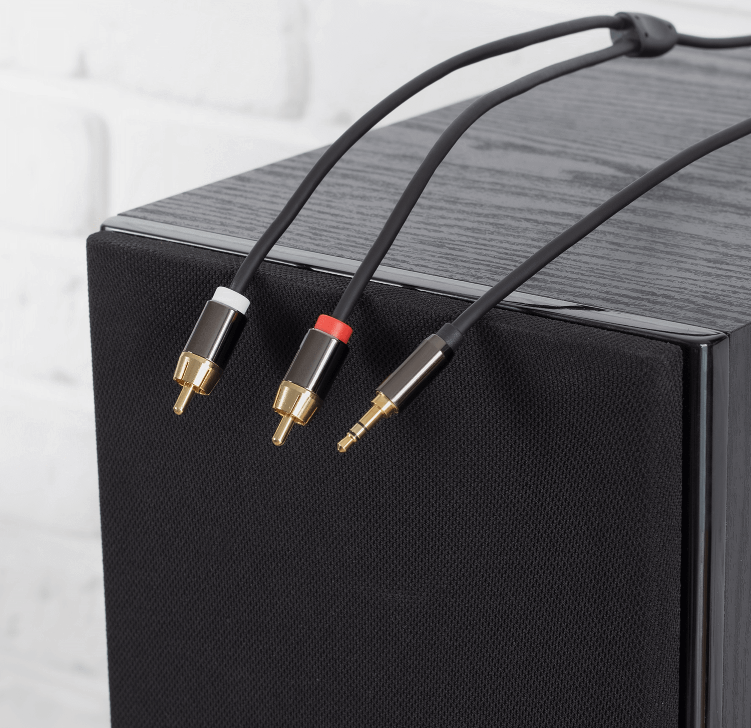 Na obrazie widoczne są trzy kable audio: dwa z końcówkami RCA w kolorach białym i czerwonym oraz jeden z końcówką Jack 3.5 mm, wszystkie z pozłacanymi wtykami. Kable leżą na czarnej powierzchni głośnika z drewnianym wykończeniem.