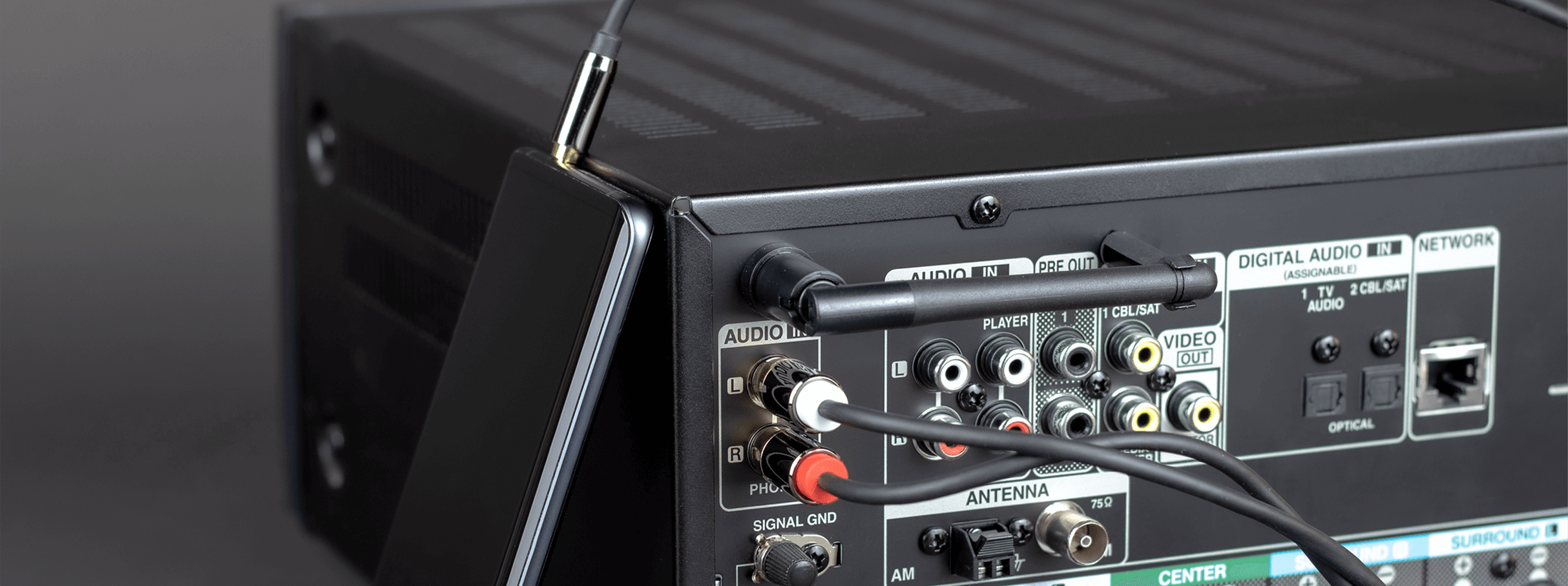 Obraz przedstawia tylny panel urządzenia audio z różnymi wejściami i wyjściami, do którego podłączony jest kabel Jack 3.5 mm - 2x RCA. Widać również smartfon podłączony do urządzenia za pomocą kabla Jack.