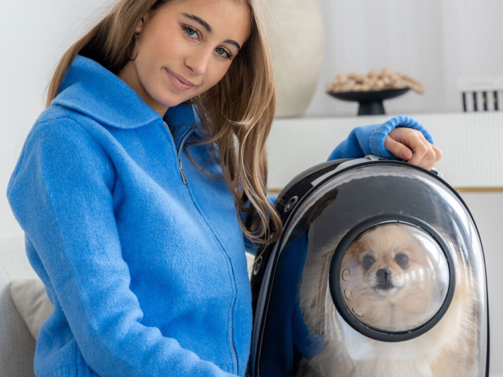 Uśmiechnięta kobieta w niebieskiej bluzce siedzi obok plecaka-nosidła, w którym siedzi mały, puszysty pies rasy pomeranian. Pies patrzy w kamerę.