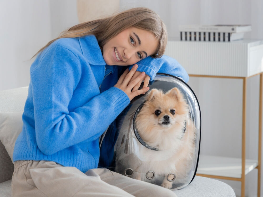 Kobieta w niebieskiej bluzce opiera się o plecak-nosidło z puszystym psem rasy pomeranian w środku. Kobieta patrzy w stronę psa, który siedzi spokojnie.