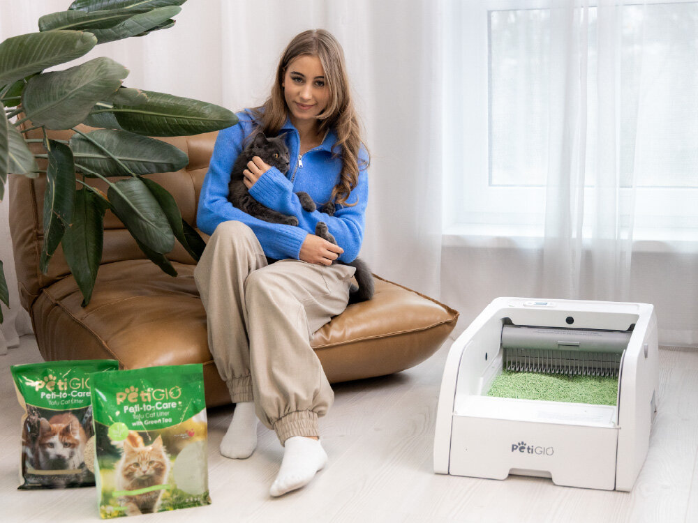 PetiGIO żwirek tofu Green Tea PETI-261 2,5 kg Kobieta w niebieskim swetrze siedzi na brązowym fotelu i trzyma na kolanach szarego kota. Obok nich, na jasnej podłodze, stoi biała inteligentna kuweta PetiGiò wypełniona zielonym żwirkiem oraz dwa opakowania żwirku tofu Peti-to-Care.