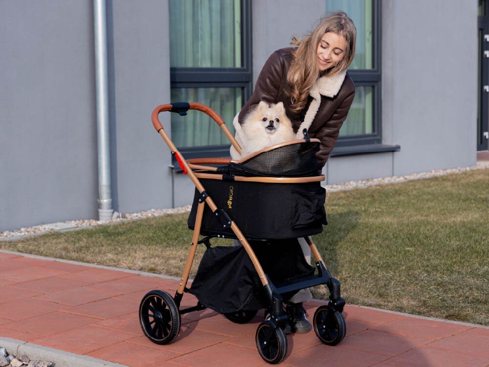 Wózek dla zwierząt PETIGIO Peti-to-Travel PETI-705-B Młoda kobieta w brązowej kurtce kożuchowej pochyla się nad czarnym wózkiem dla psa PettiGio na zewnątrz, uśmiechając się do jasnego Pomeraniana siedzącego w środku. W tle widać budynek i trawnik.