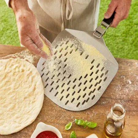 Osoba posypuje łopatkę do pizzy mąką, obok znajduje się ciasto na pizzę na drewnianym blacie. W tle widoczne są zielone liście bazylii i mała butelka z oliwą.
