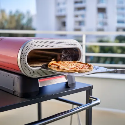 Na obrazie widoczny jest piec do pizzy z otwartymi drzwiczkami, z którego wyciągana jest pizza na metalowej łopatce. W tle znajdują się rozmyte budynki i balustrada.