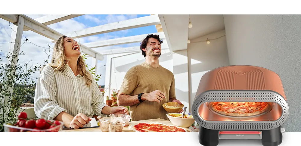 Na obrazie widać dwie osoby stojące przy stole z jedzeniem na zewnątrz oraz piec do pizzy z widoczną pizzą w środku. Na piecu znajduje się logo \'tefal\'.