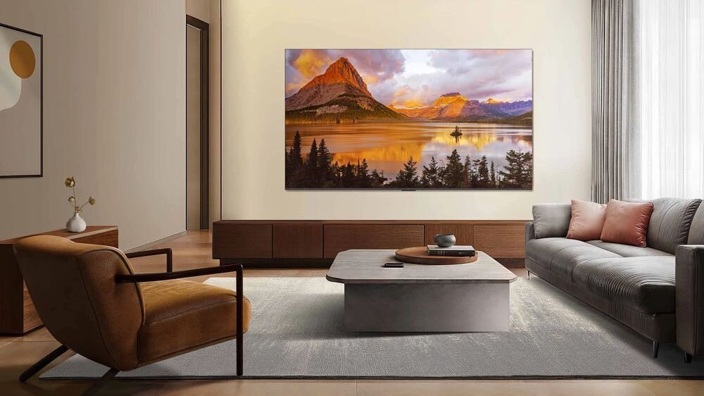 W elegancko urządzonym salonie na ścianie wisi duży telewizor wyświetlający krajobraz górski z jeziorem. W pomieszczeniu znajdują się także sofa, fotel i stolik kawowy.