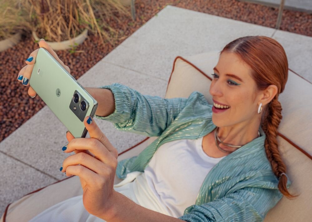 Osoba trzyma smartfon w kolorze zielonym z widocznym logo producenta na tylnej obudowie. W tle widać rośliny i fragment tarasu.