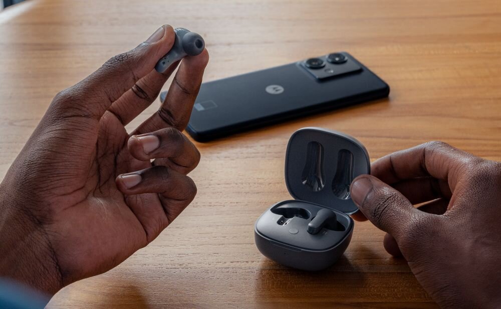 Na drewnianym stole leży czarny smartfon z logo w kształcie litery \'M\'. Obok osoba trzyma szare słuchawki bezprzewodowe i otwarte etui ładujące.