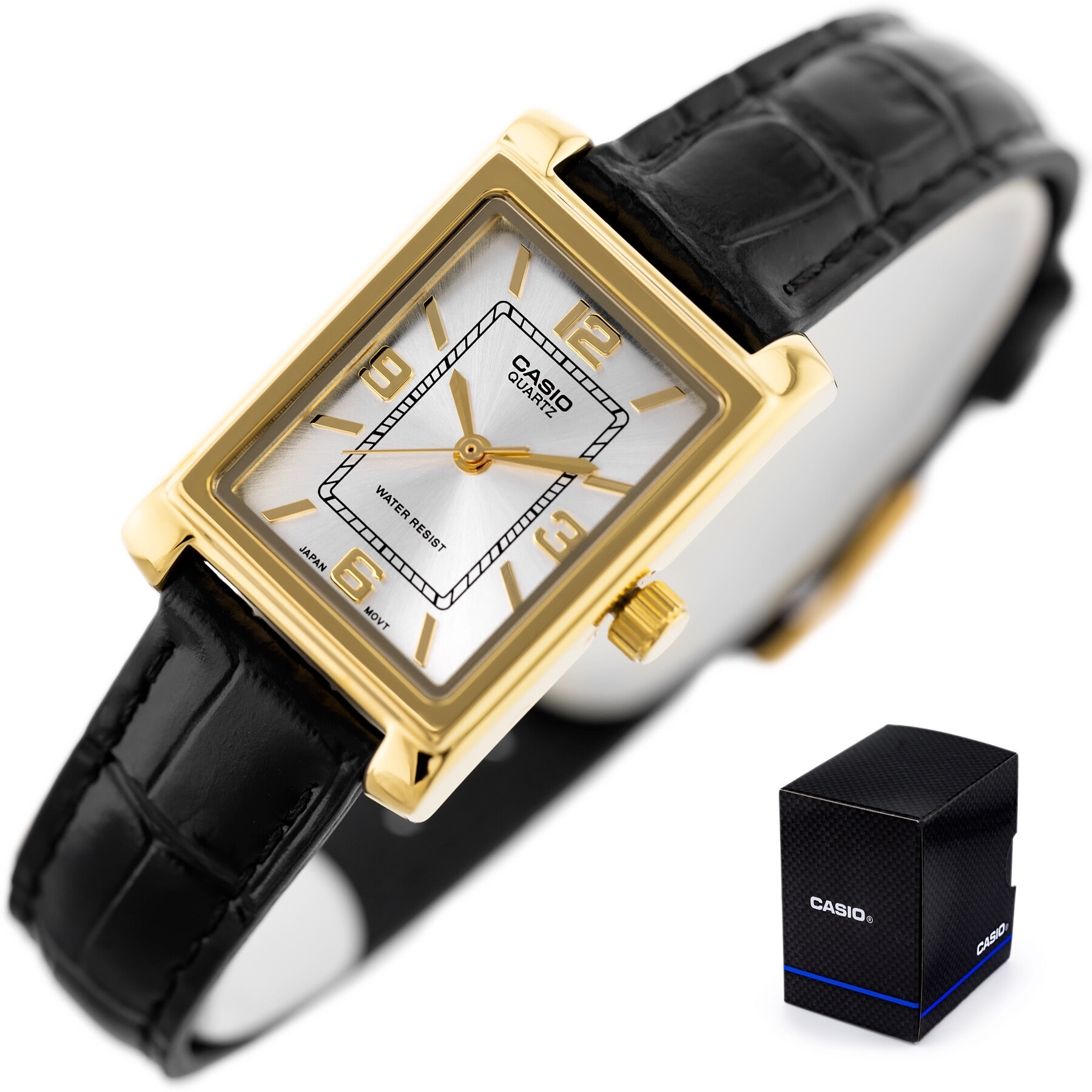 Zegarek z prostokątną, złotą kopertą i białą tarczą z oznaczeniami godzinowymi oraz czarnym paskiem. Obok znajduje się czarne pudełko z napisem \'CASIO\'.