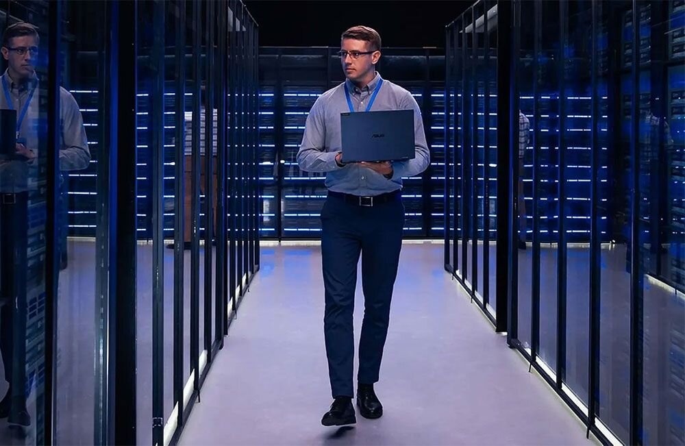 Osoba trzymająca laptopa przechodzi przez korytarz serwerowni z rzędem szaf serwerowych po obu stronach. Na laptopie widoczne jest logo ASUS.