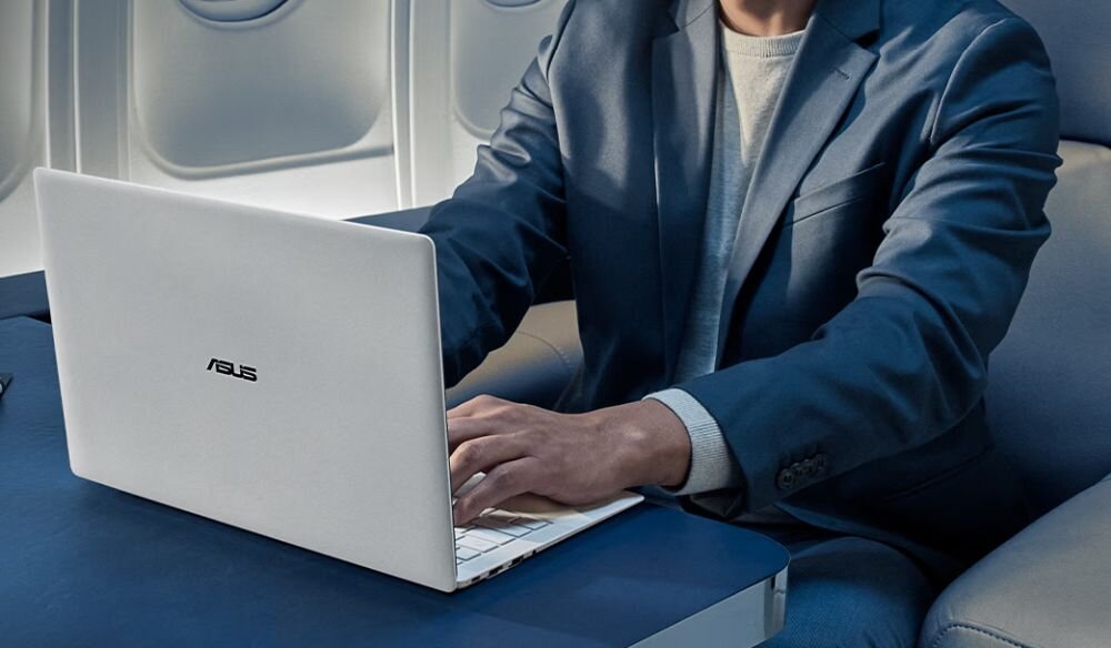Osoba w garniturze korzysta z białego laptopa z logo ASUS, siedząc przy stoliku w samolocie.