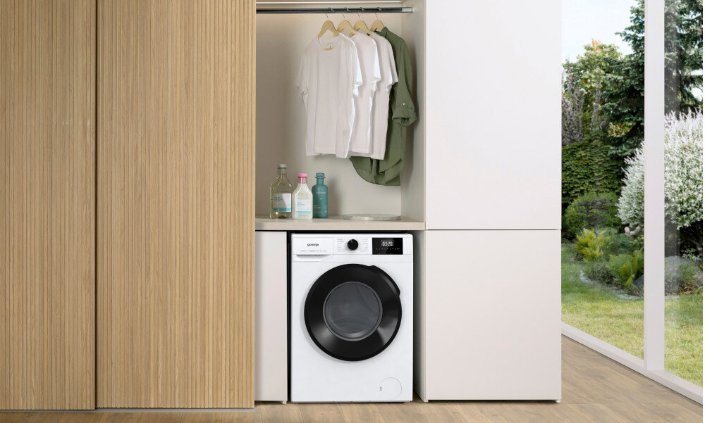 PRALKA GORENJE W3NGPI61SAS Zdjęcie przedstawia pralkę w zabudowie pod półką z powieszonymi ubraniami i akcesoriami do prania. Całość pokazuje praktyczne wykorzystanie przestrzeni oraz uporządkowane miejsce na domowe tekstylia