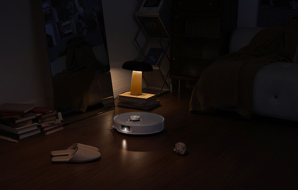 Na podłodze w ciemnym pokoju znajduje się biały robot sprzątający, obok którego leżą kapcie i zmięta kartka papieru. W tle widać lampkę stołową emitującą ciepłe światło oraz stos książek.
