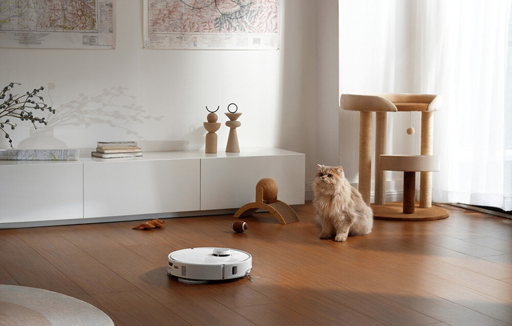 W jasnym pokoju na drewnianej podłodze znajduje się biały robot sprzątający. Obok niego siedzi kot, a w tle widać białą szafkę i drapak dla kota.