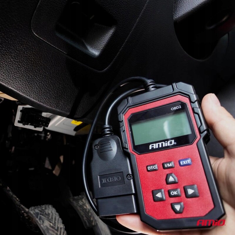 Osoba trzyma urządzenie diagnostyczne AMiO OBD2 z wyświetlaczem LCD i przyciskami oznaczonymi jako DTC, I/M, EXIT, OK oraz strzałkami. W tle widoczna jest część wnętrza samochodu z gniazdem OBD2.