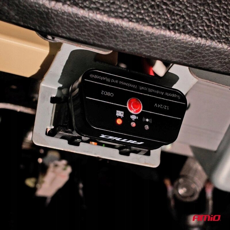 Na zdjęciu widoczny jest interfejs diagnostyczny AMiO OBD2 podłączony do gniazda w samochodzie. Na urządzeniu znajdują się napisy: \'Supports Android/iOS/Windows and Bluetooth\', \'12/24V\', \'OBD2\', oraz logo \'AMiO\'.