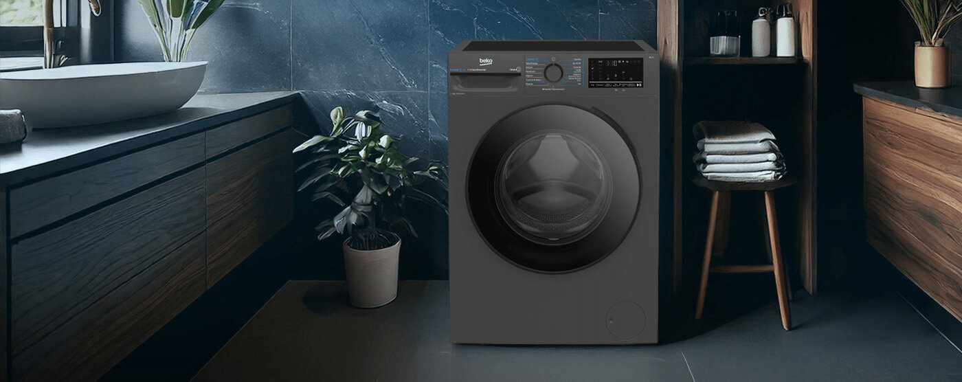 Pralka w kolorze ciemnym stoi w nowoczesnej łazience z ciemnymi ścianami i drewnianymi szafkami. Obok pralki znajduje się roślina w doniczce oraz półka z ręcznikami i kosmetykami.