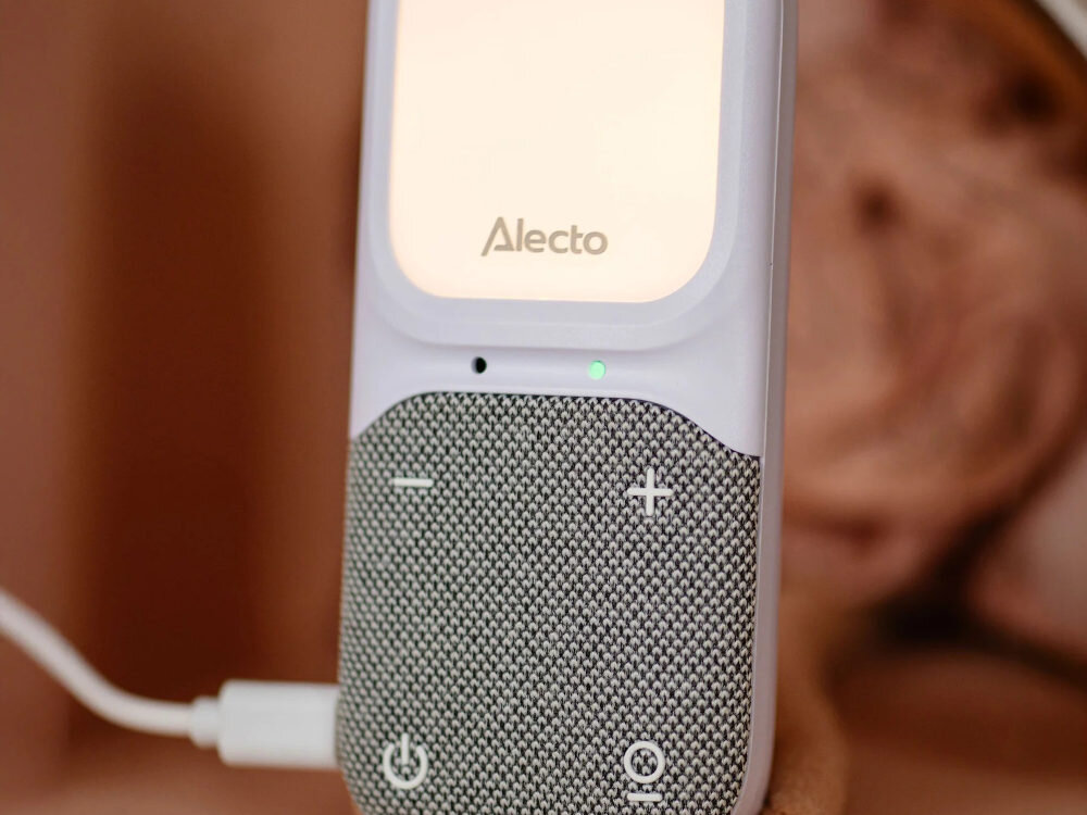 Na zdjęciu widoczna jest elektroniczna niania z logo \'Alecto\'. Urządzenie ma teksturowaną powierzchnię z przyciskami plus i minus oraz ikonami zasilania i dźwięku.