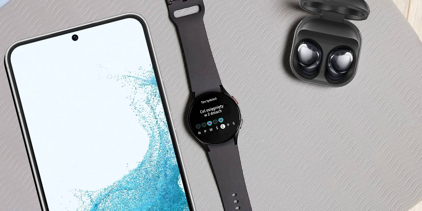 Na stole leży smartfon z wyświetlaczem włączonym, obok znajduje się czarny smartwatch z widocznym ekranem oraz otwarte etui z dwoma słuchawkami. Na ekranie smartwatcha widoczny jest tekst: \'Cel osiągnięty w 2 dniach\'.
