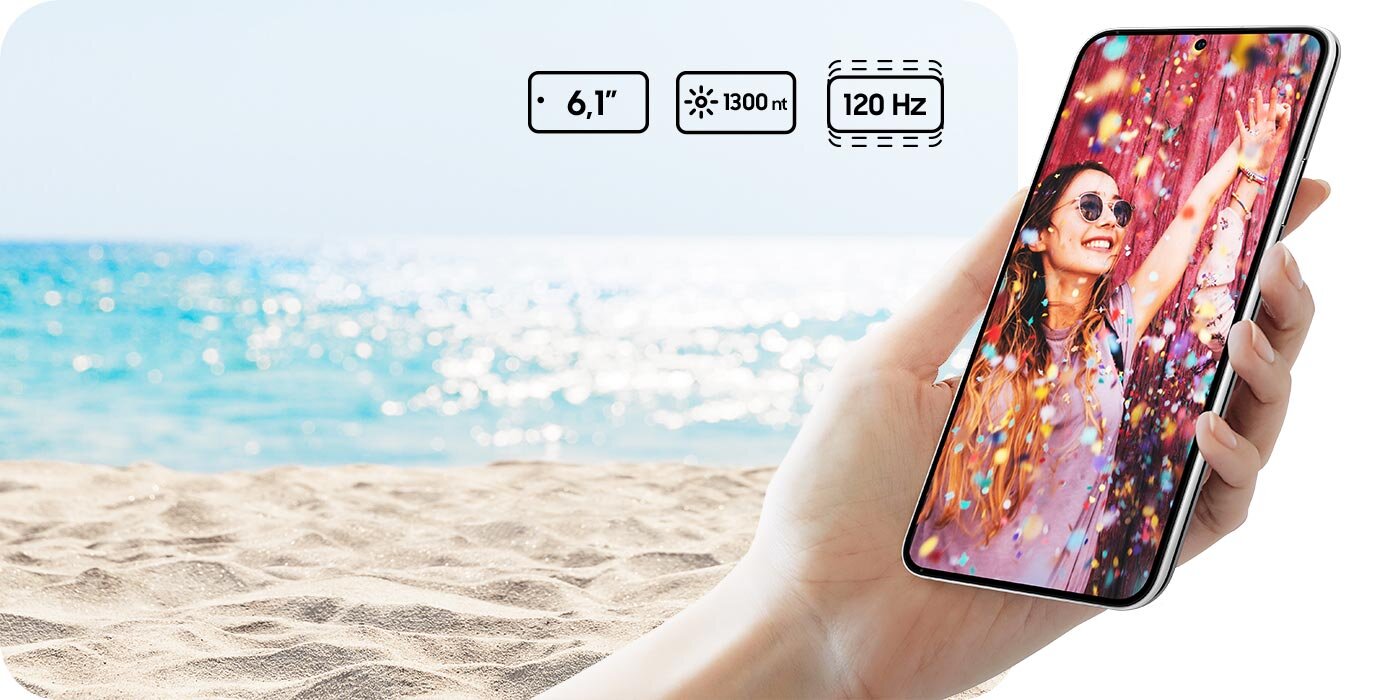 Osoba trzyma smartfon z kolorowym wyświetlaczem, na którym widać uśmiechniętą kobietę w okularach przeciwsłonecznych. W tle znajduje się plaża i morze, a na obrazie widoczne są ikony z napisami: \'6.1\