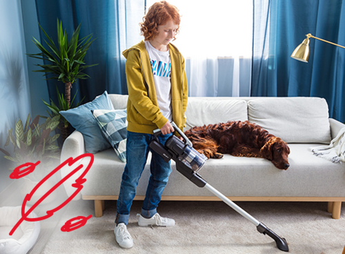 Osoba używa odkurzacza pionowego w salonie, obok na kanapie leży pies. W tle widoczne są zasłony, roślina doniczkowa i lampa stojąca.