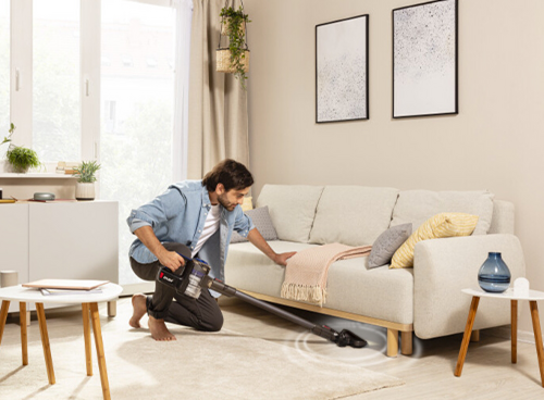 Osoba używa odkurzacza pionowego w salonie z jasną sofą i dwoma obrazami na ścianie. Na stoliku obok znajduje się niebieski wazon.