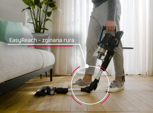 Osoba używa odkurzacza z funkcją \'EasyReach - zginana rura\' na drewnianej podłodze obok kanapy. W tle widoczne są zasłony i roślina w doniczce.
