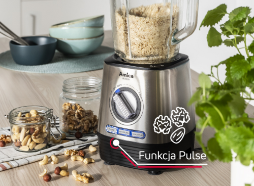Na blacie kuchennym stoi blender kielichowy marki Amica z pokrętłem i napisem \'Funkcja Pulse\'. Obok znajdują się słoiki z orzechami i miski, a w tle widoczna jest roślina.