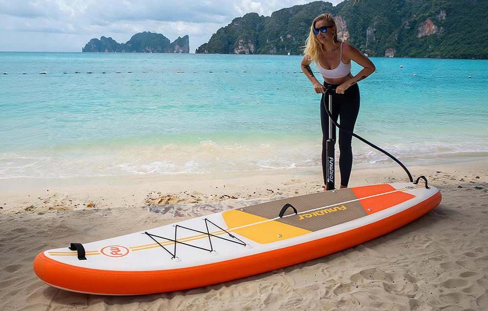 Deska SUP FUNWATER SUPFW69B Pomarańczowo-biały, uniwersalna, deska, rekreacja, jezioro, Kobieta pompuje deskę SUP na piaszczystej plaży.