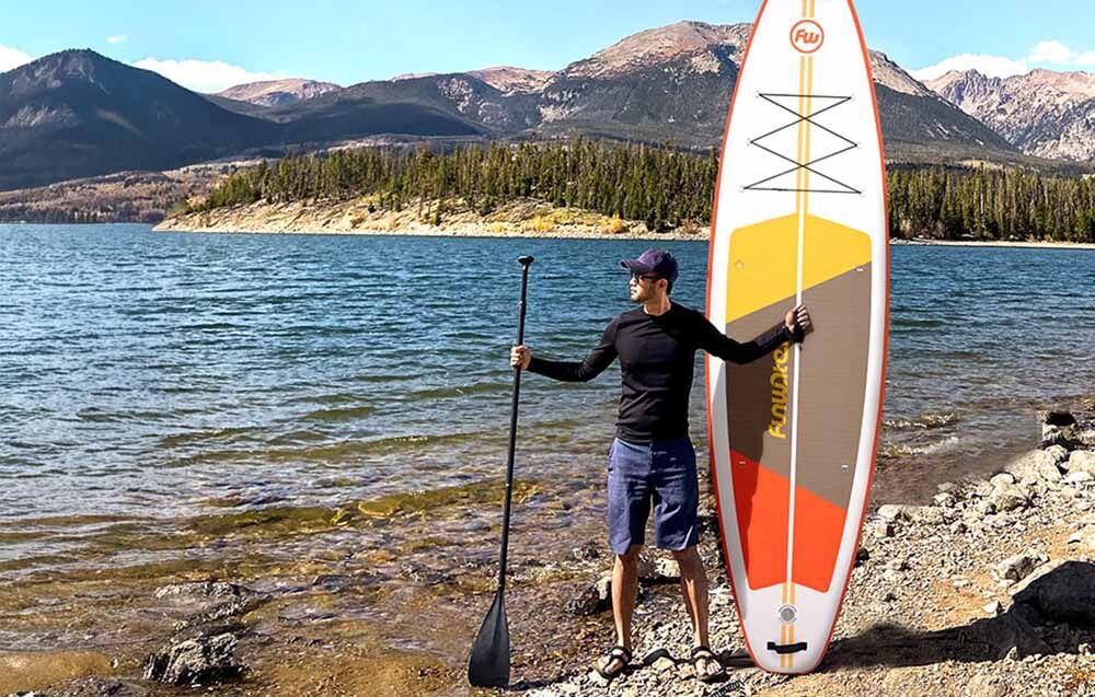 Deska SUP FUNWATER SUPFW69B Pomarańczowo-biały, pianka, EVA, przyczepność, joga, Mężczyzna stoi z wiosłem obok deski nad jeziorem.