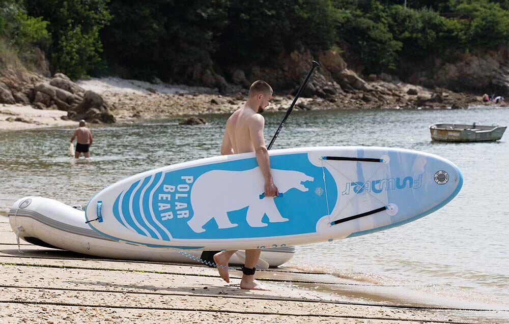 Deska SUP FUNWATER SUPFW07A Niebieski, uniwersalna, deska, rekreacja, jezioro, Mężczyzna niesie biało-niebieską deskę SUP z grafiką niedźwiedzia polarnego brzegiem kamienistej plaży.