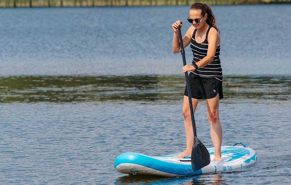 Deska SUP FUNWATER SUPFW07A Niebieski, uniwersalna, deska, rekreacja, jezioro, Kobieta w okularach przeciwsłonecznych płynie na stojąco deską SUP, trzymając czarne wiosło nad taflą wody.