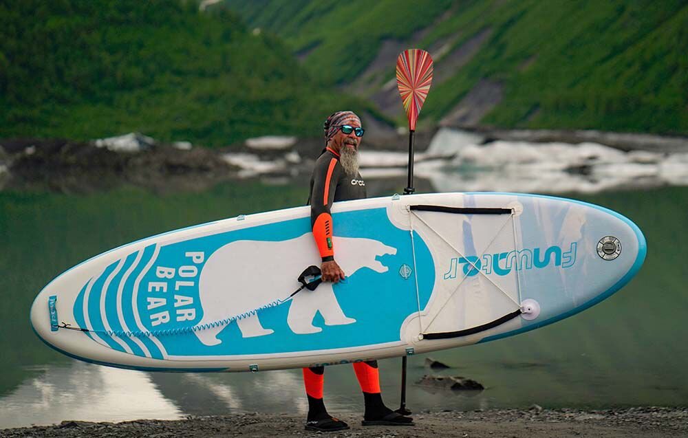 Deska SUP FUNWATER SUPFW07A Niebieski, stabilność, manewrowanie, stateczniki, kontrola, Brodaty mężczyzna w piance trzyma deskę SUP oraz kolorowe wiosło na tle górskiego jeziora i lodowca.