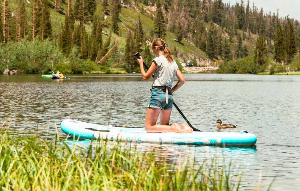 Deska SUP FUNWATER SUPFW07A Niebieski, pianka, EVA, przyczepność, joga, Dziewczyna w jeansowych szortach wiosłuje klęcząc na desce SUP. W pobliżu po tafli jeziora pływa kaczka