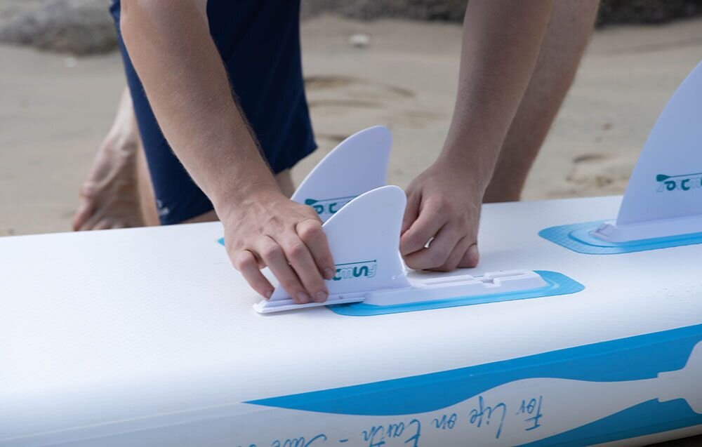 Deska SUP FUNWATER SUPFW07A Niebieski, uchwyt, manometr, linki, sztywność, Zbliżenie na dłonie montujące biały statecznik w gnieździe na spodzie deski SUP leżącej na piasku.