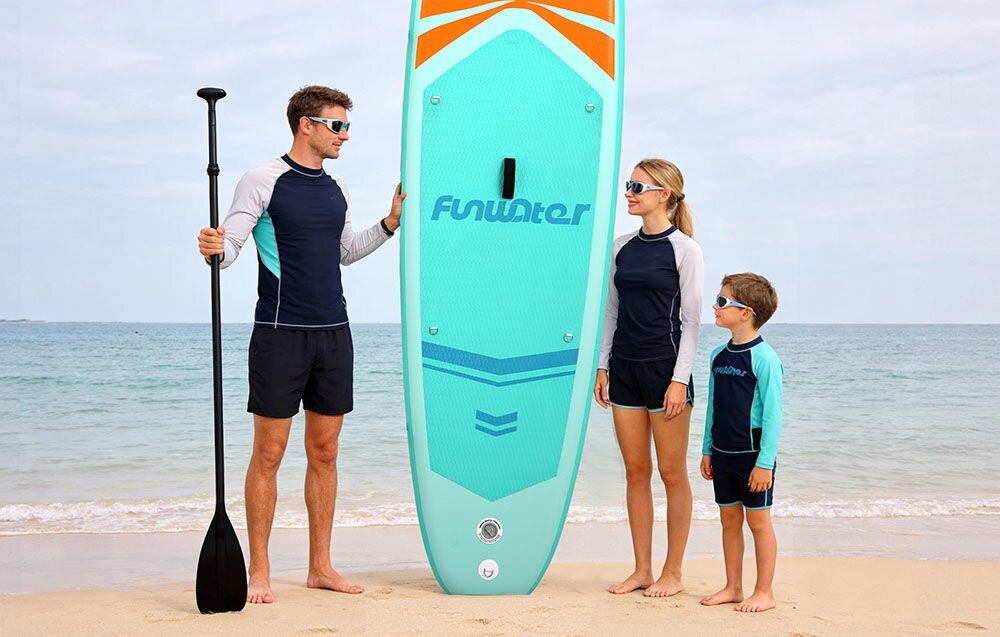 Deska SUP FUNWATER SUPFW70A Turkusowy, pianka, EVA, przyczepność, joga, Kobieta niesie deskę SUP po piaszczystej plaży.