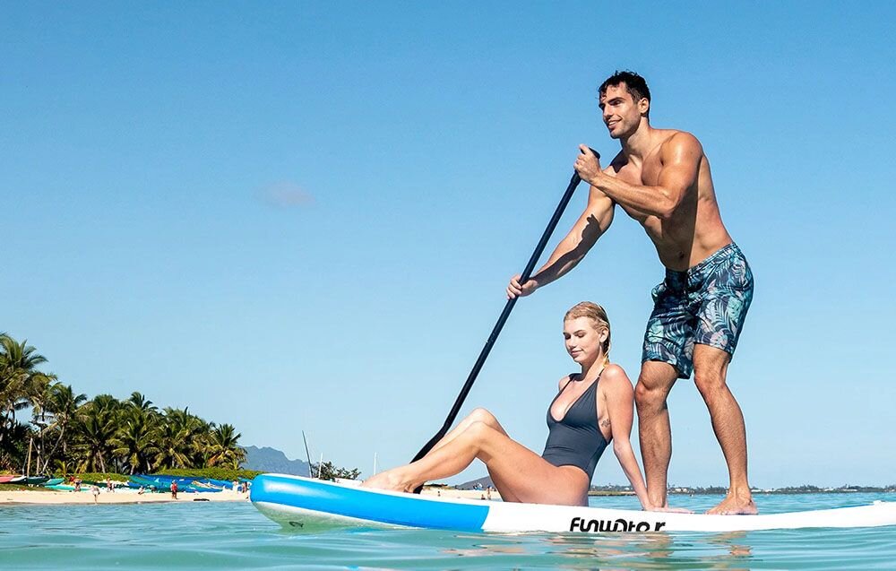 Deska SUP FUNWATER SUPFW05B Niebiesko-biały, pianka, EVA, przyczepność, joga, Kobieta niesie deskę SUP po piaszczystej plaży.