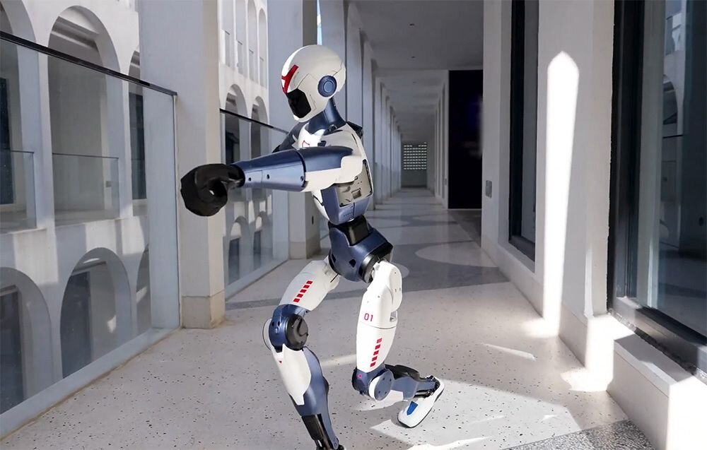 Robot humanoidalny w białym i niebieskim kolorze z czerwonymi akcentami, wykonujący ruch w korytarzu z przeszklonymi ścianami. Robot ma zgięte ramiona i nogi, a jego powierzchnia jest gładka i nowoczesna.
