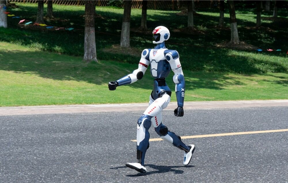 Robot humanoidalny o futurystycznym wyglądzie idzie po asfaltowej drodze. W tle widoczne są drzewa i trawnik.