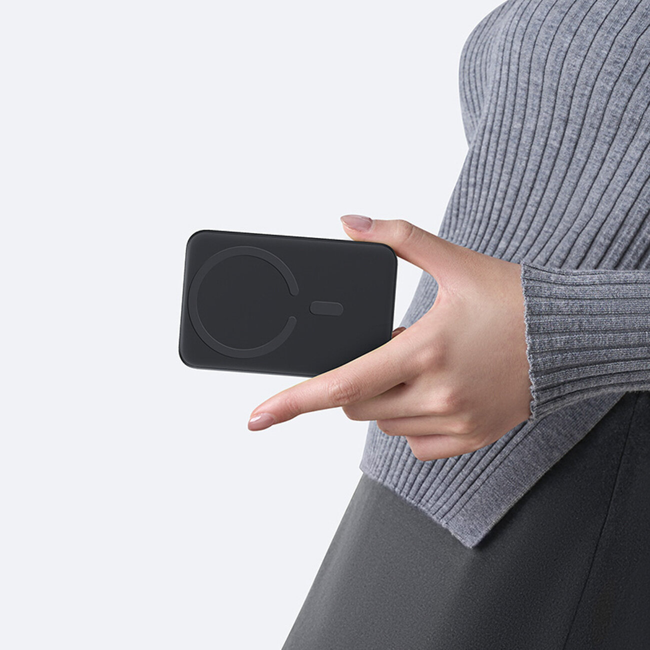 Osoba trzyma czarny power bank z okrągłym wzorem na powierzchni. Ubrana jest w szary sweter i ciemne spodnie.