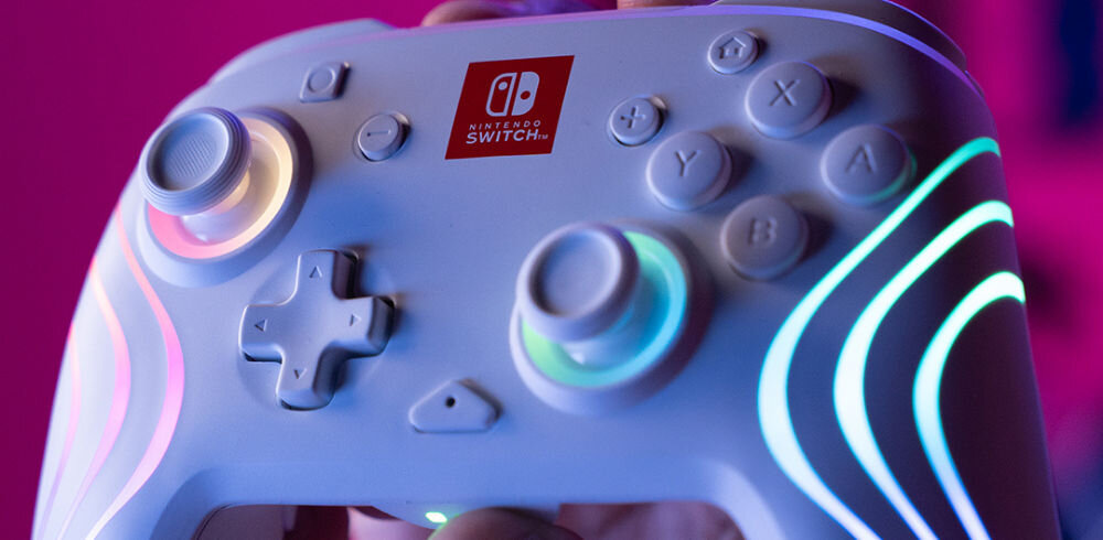 Kontroler PDP Afterglow Wave Nintendo Switch/Nintendo Switch Oled Biały prezentacja kontrolera trzymanego w ręce na różowym tle podświetlonego pokaz świetlny 8 niezależnych stref RGB 4 gotowe tryby graj w rytmie światła