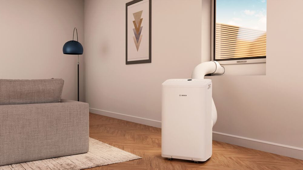 W jasnym pokoju znajduje się biały klimatyzator z logo BOSCH, podłączony rurą do otwartego okna. Obok stoi szara sofa i lampa, a na ścianie wisi obraz z geometrycznym wzorem.