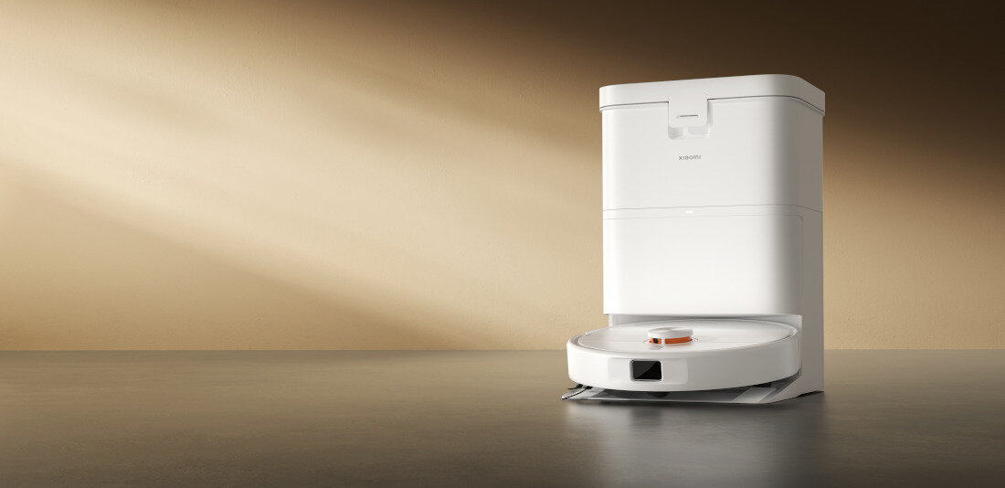 Na obrazie znajduje się biały robot sprzątający z bazą ładującą na tle brązowej ściany. Na urządzeniu widoczne jest logo \'XIAOMI\'.