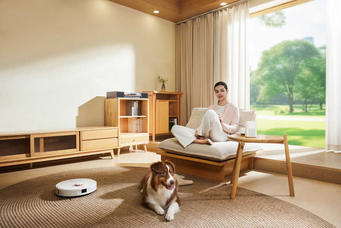 W jasnym, nowoczesnym salonie na dywanie znajduje się biały robot sprzątający. Na sofie siedzi osoba, a obok leży pies.